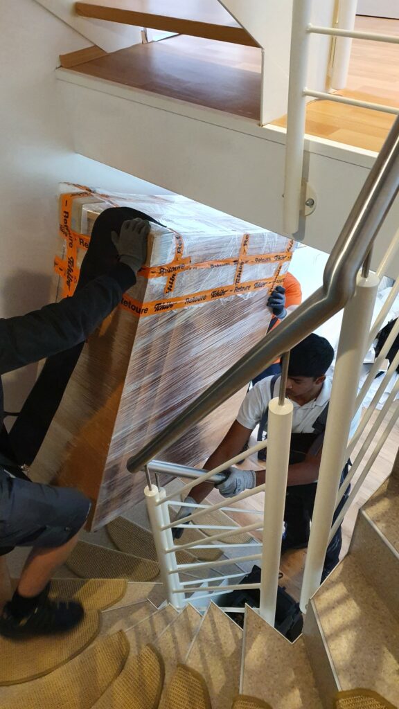 Zwei Mitarbeiter tragen ein schweres Möbelstück über eine Wendeltreppe herunter